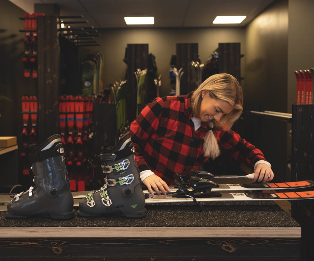 A person in a red plaid shirt adjusts ski bindings in a shop, with ski boots and skis visible on the counter.