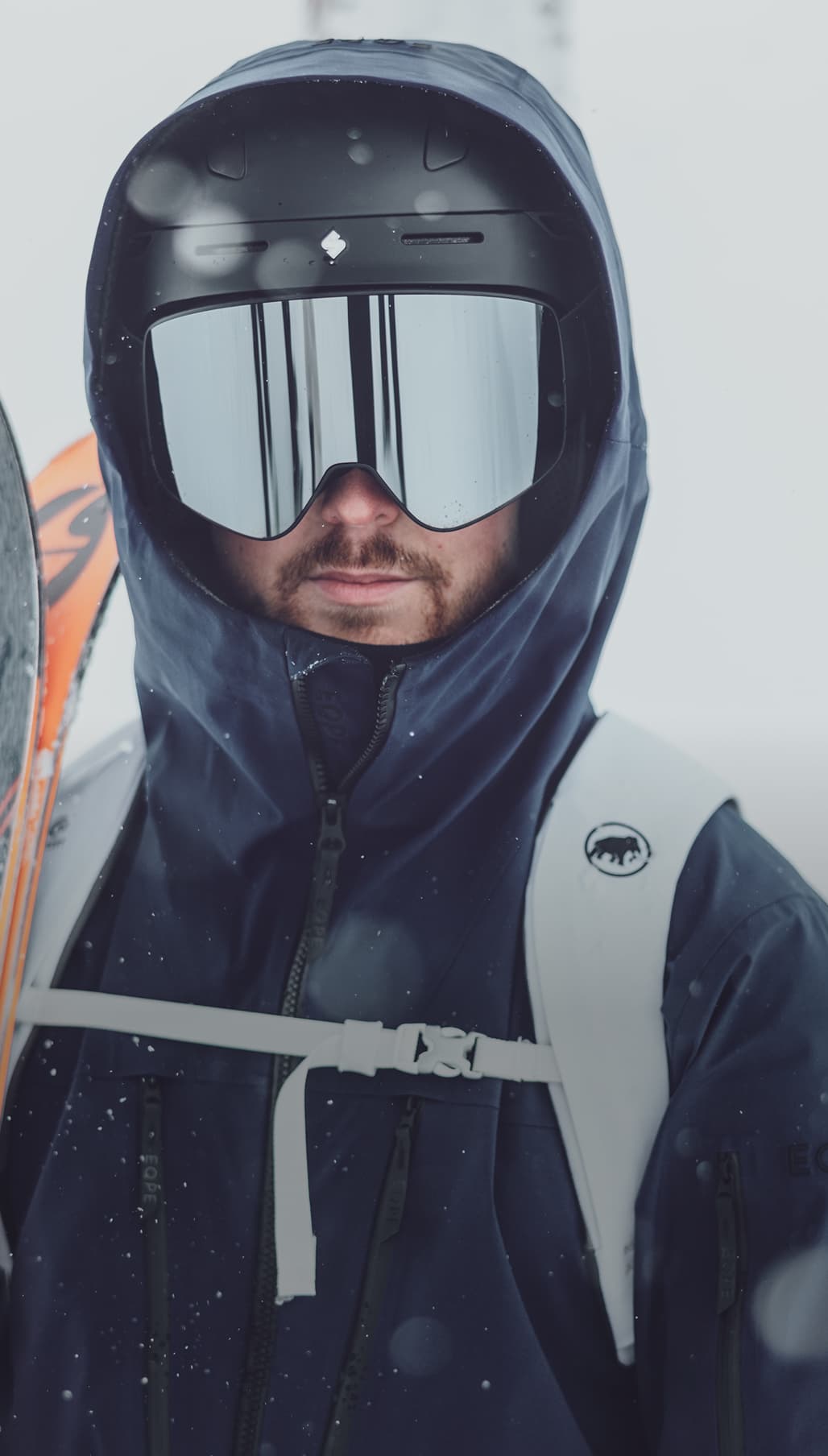 Person in a dark blue ski jacket and helmet with reflective goggles, holding skis, surrounded by falling snow.