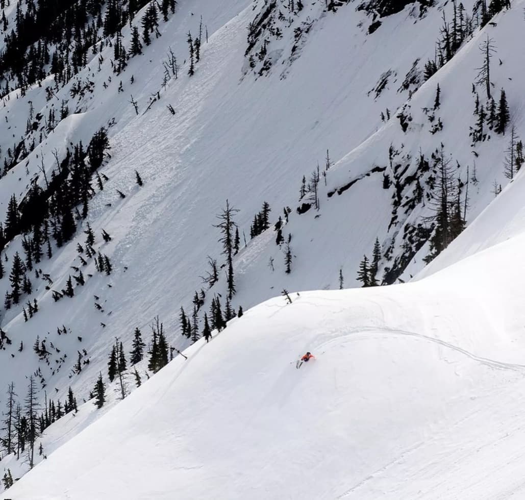 Snowy mountain slope with scattered trees and a skier in red descending, leaving a trail in the snow.