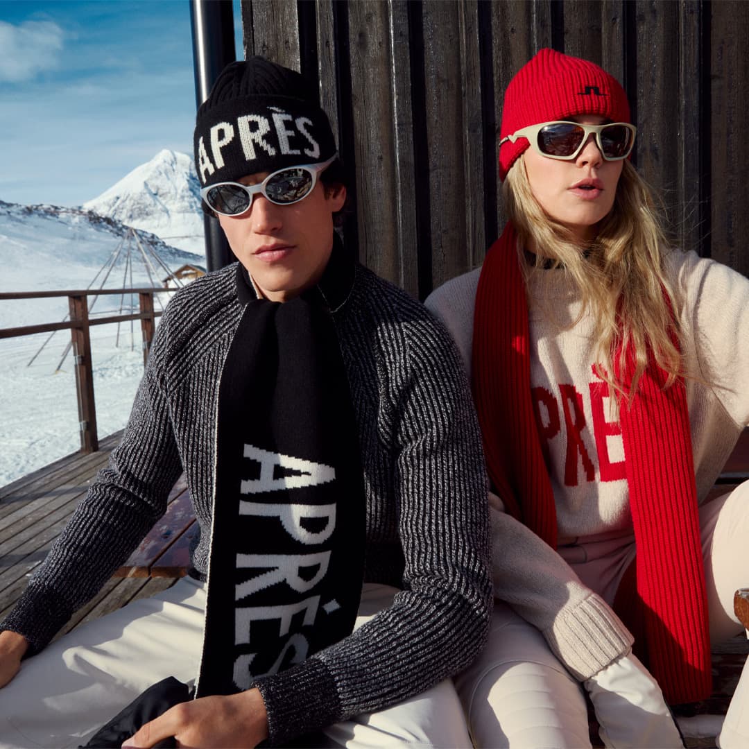 Two people in winter attire, wearing hats, sunglasses, and scarves, sit outside a wooden cabin with a snowy mountain in the background.