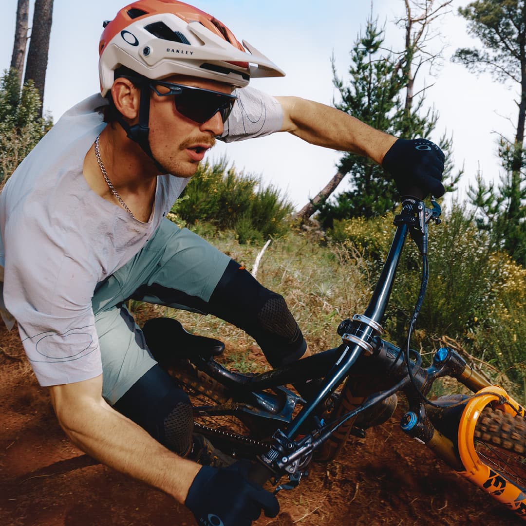 Mountain biker in a helmet and sunglasses navigating a dirt trail through a forest, focused and leaning into a turn.