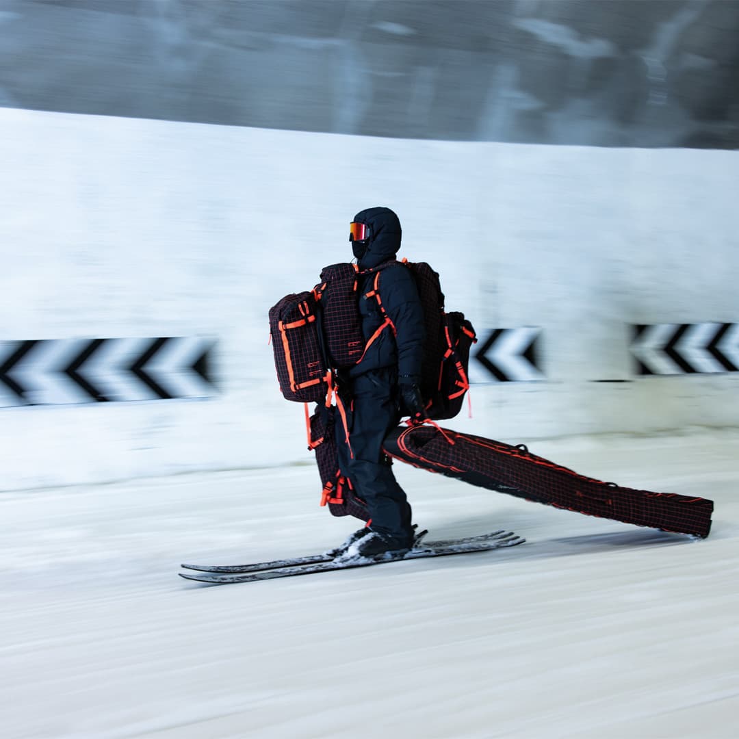 Person skiing indoors, wearing a black and red suit with large, rectangular backpacks, against a blurred background with directional arrows.