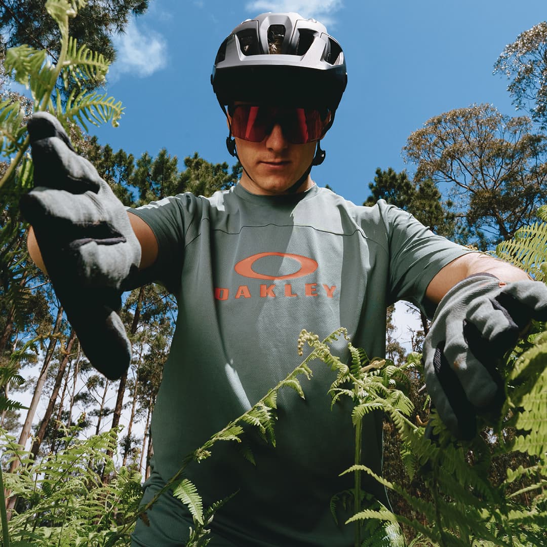 A cyclist wearing a helmet, sunglasses, and gloves stands among green ferns, against a backdrop of trees and a clear blue sky.