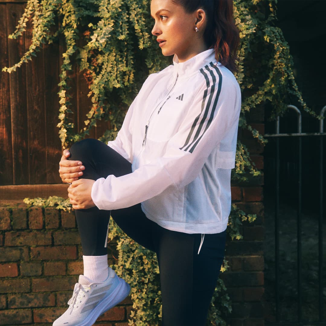 A woman in a athletic gear stretches her leg outdoors, surrounded by greenery and brickwork.