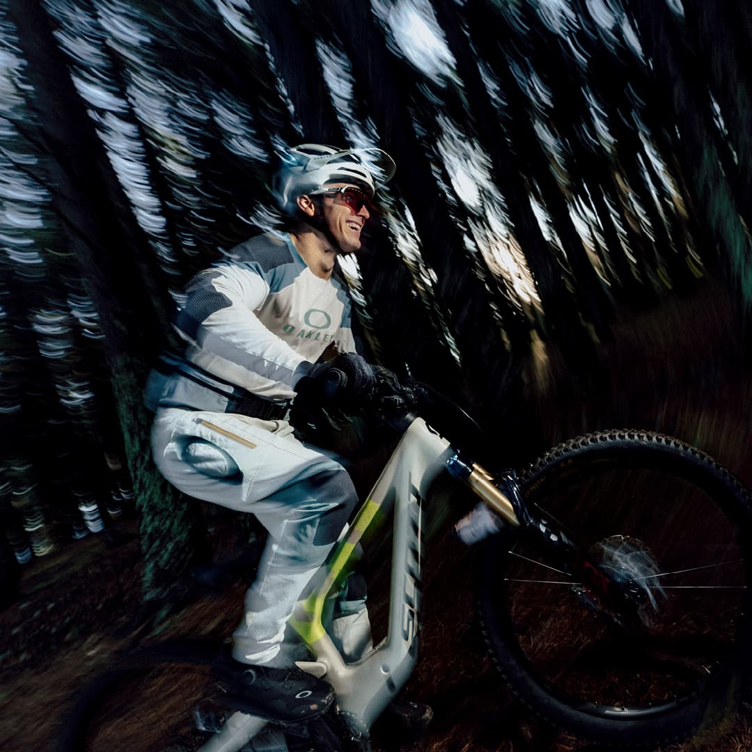 A cyclist wearing a helmet and gear rides a mountain bike through a forest, with motion blur emphasizing speed and movement.