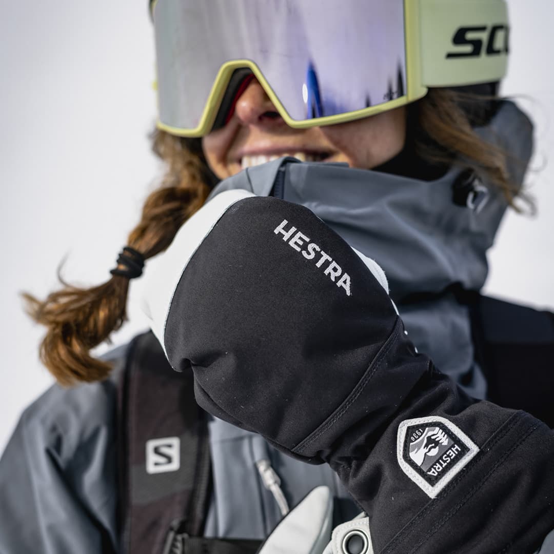 Person wearing ski gear and goggles, smiling. Close-up of black Hestra glove with logo.