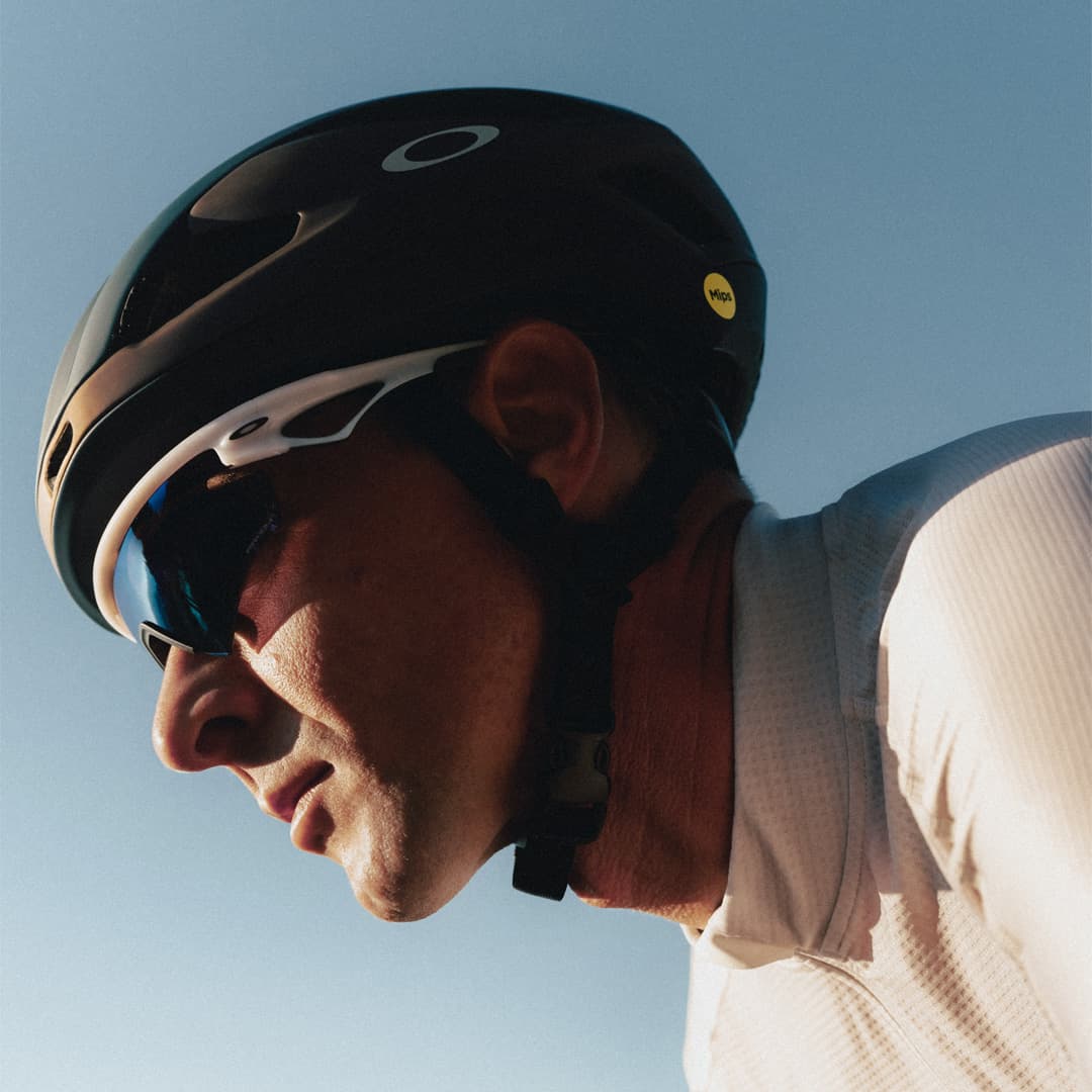 Cyclist wearing a black helmet and sunglasses, dressed in a white jersey, riding under a clear blue sky.