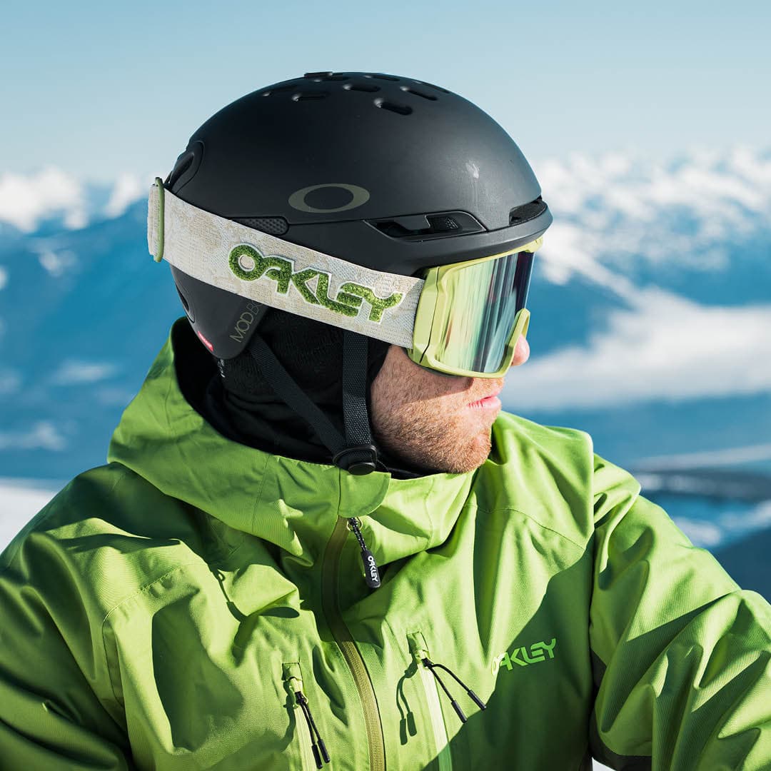 Person in a green jacket and black helmet with goggles, overlooking snowy mountains.