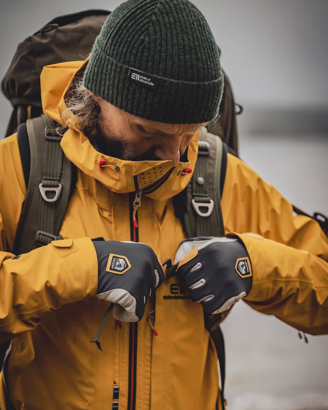 Person in a yellow jacket and green beanie adjusts their gloves, wearing a backpack, outdoors on a cloudy day.