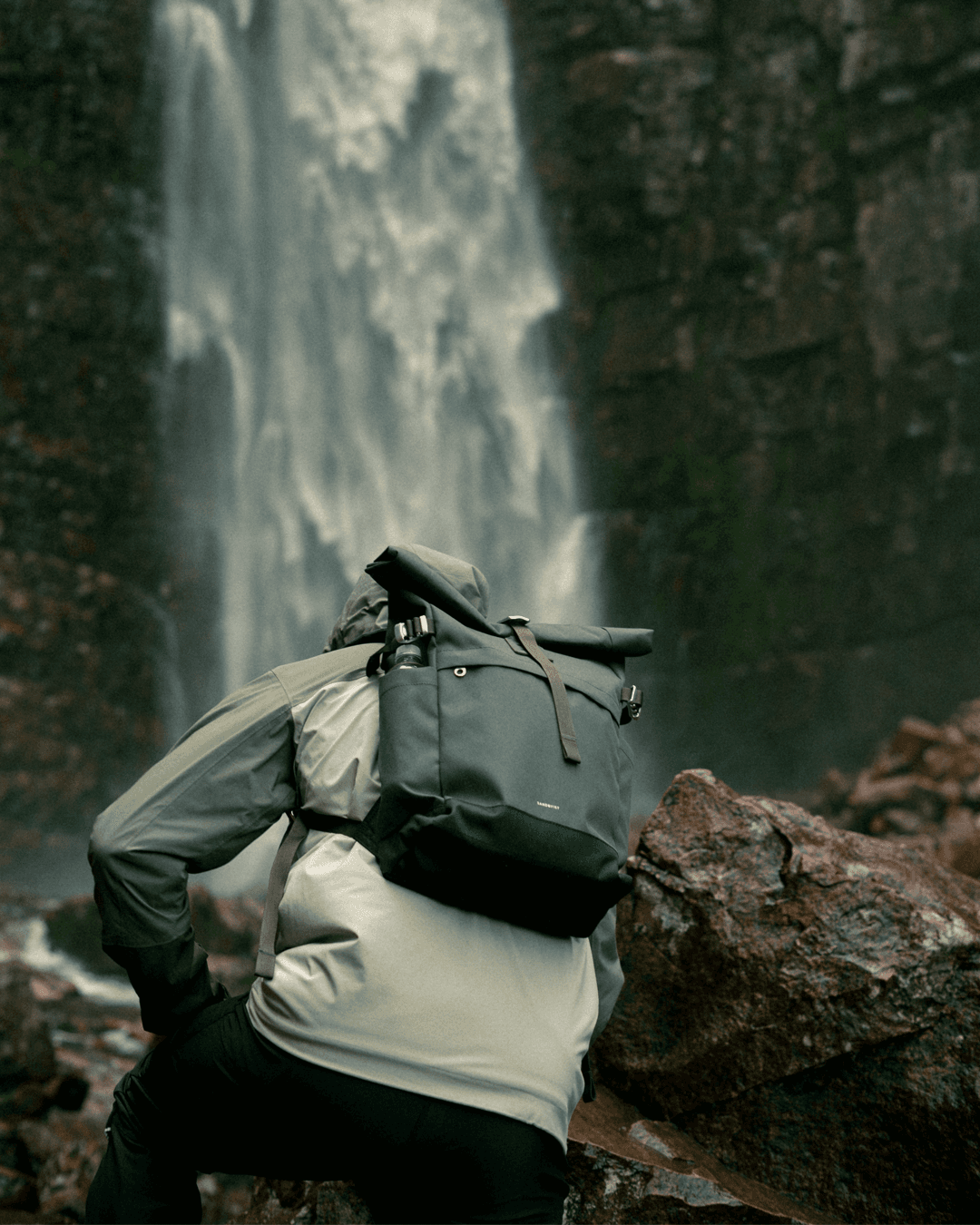 A person in a green jacket and backpack climbs over wet rocks near a waterfall in a lush, forested area.