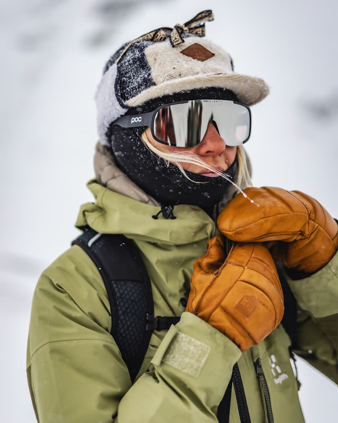 A person in a green jacket and brown mittens adjusts their ski goggles in a snowy landscape.