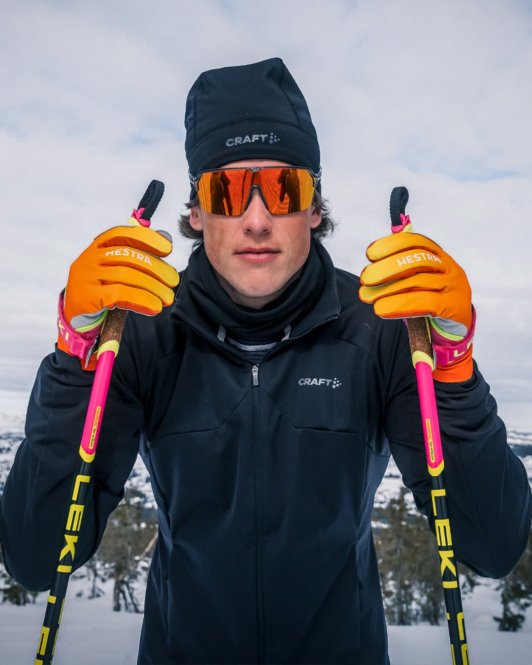 A skier in a black outfit, orange gloves, and reflective sunglasses holds ski poles against a snowy mountain backdrop.