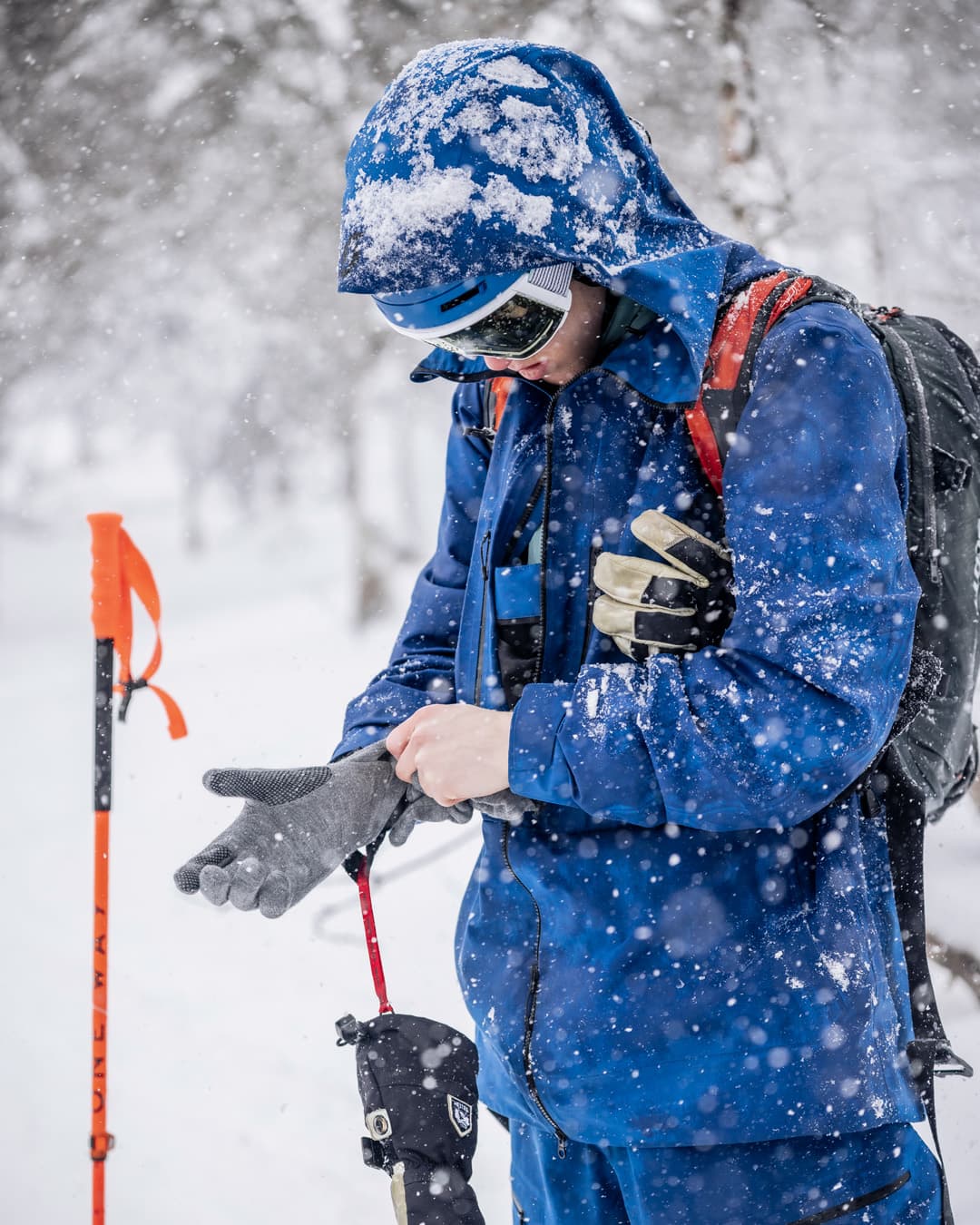 Person in blue winter gear adjusts gloves in a snowy landscape, holding ski poles, with snowflakes falling around them.