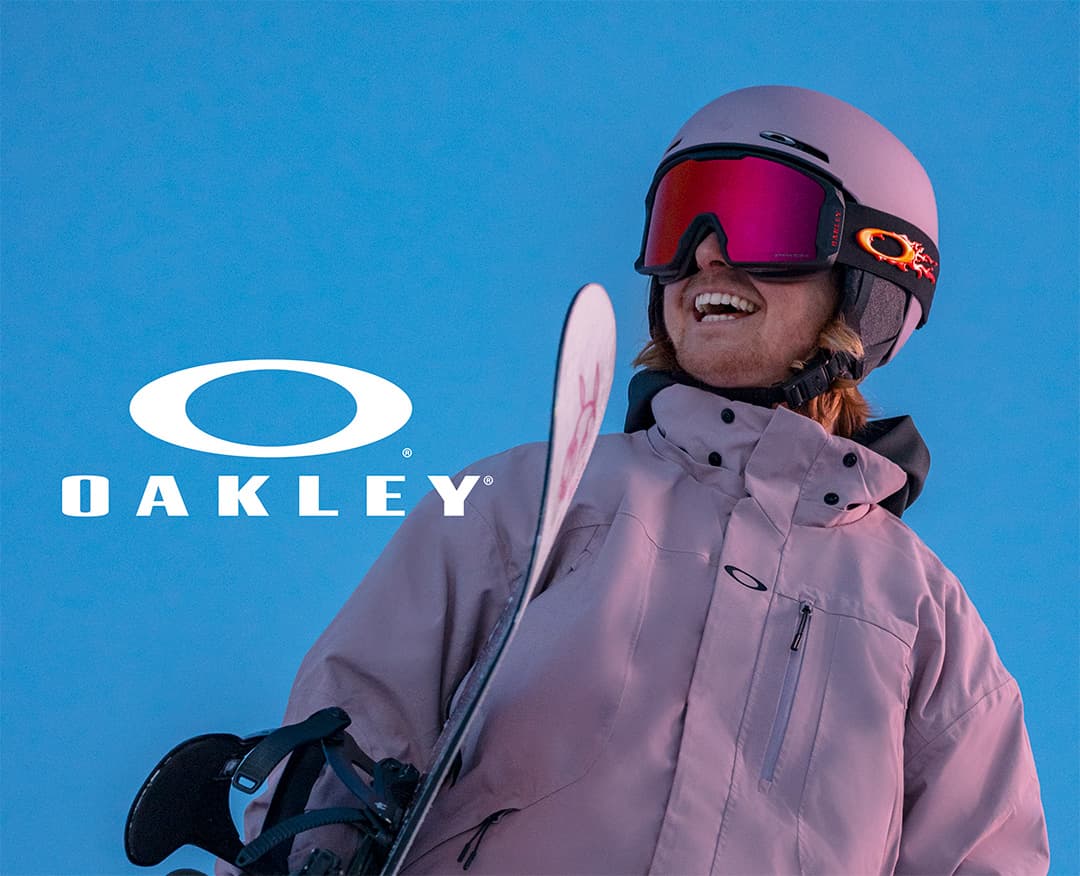 Person in a pink jacket and helmet holding a snowboard, wearing Oakley goggles, smiling against a blue sky. Oakley logo is visible.