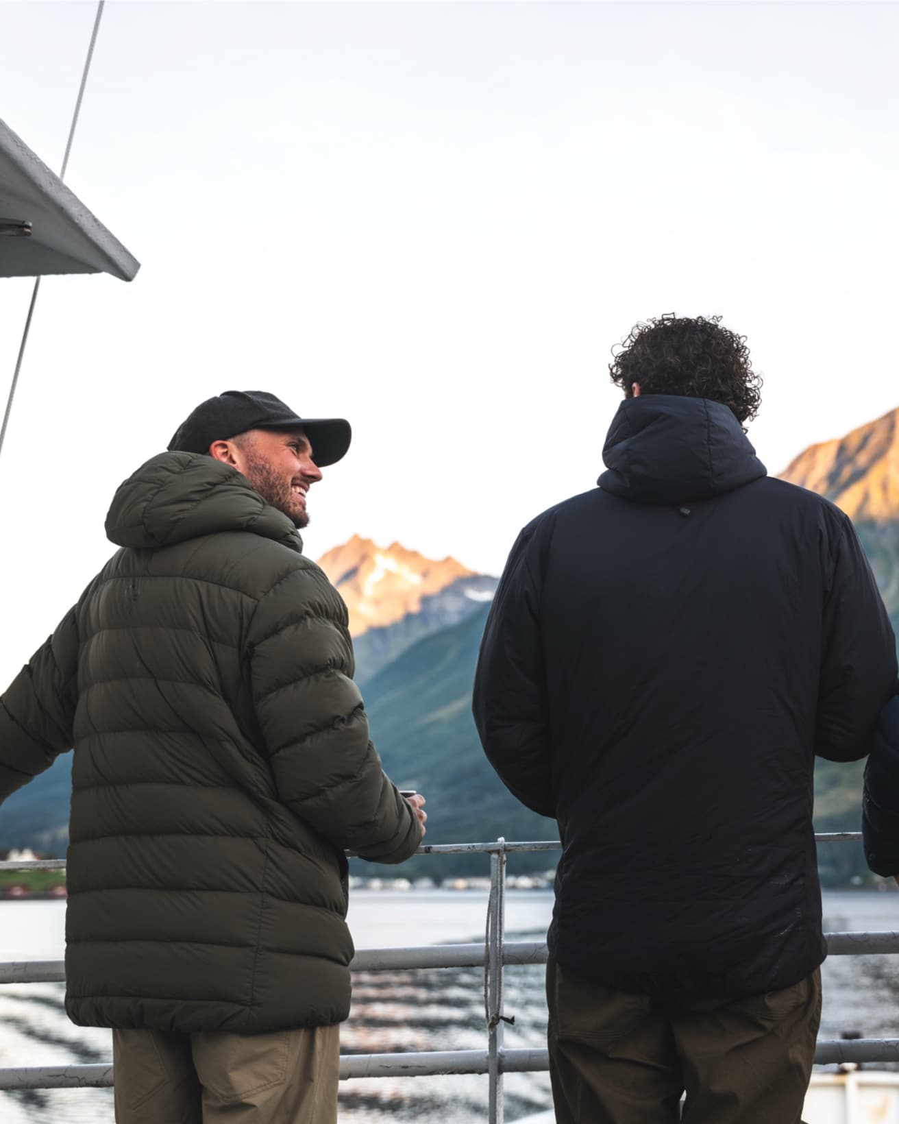 Two people in jackets on a boat, facing snowy mountain peaks in the distance, under a clear sky.