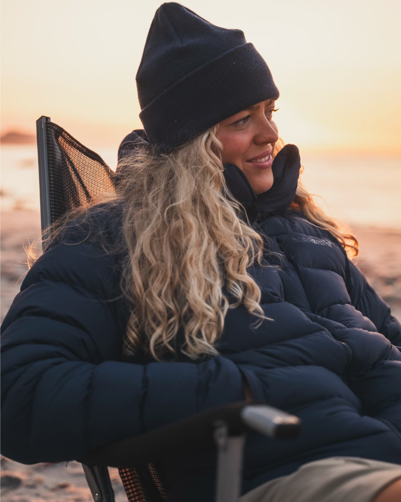 Woman with long curly hair in a black beanie and puffer jacket sits on a beach chair, smiling at sunset.