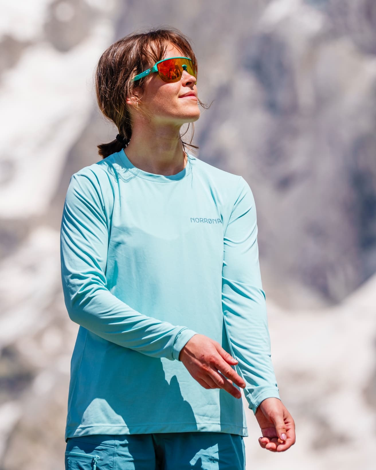 Person in a light blue long-sleeve shirt and sunglasses enjoying the outdoors, standing against a mountainous background.