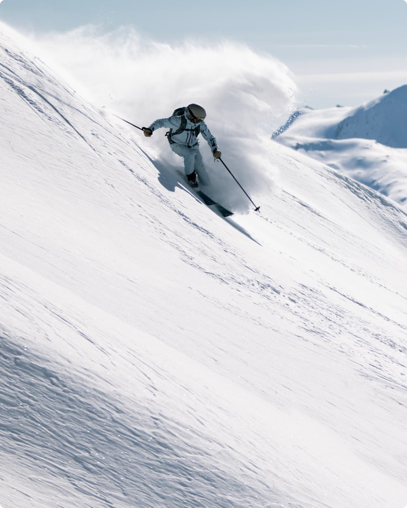 Skier in blue gear carving down a snowy slope, creating a spray of snow, with clear skies and distant mountains in the background.