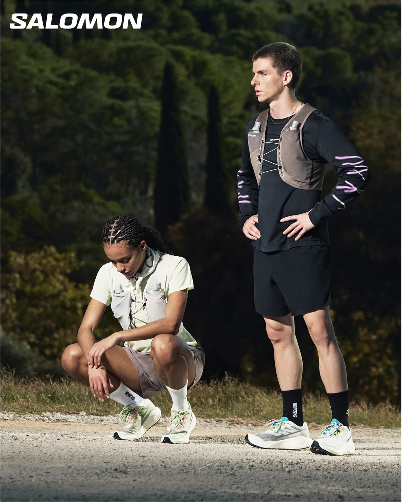 Two people in athletic gear on a trail, one standing, one crouching, both wearing running apparel. With Salomon branding in the top left corner.