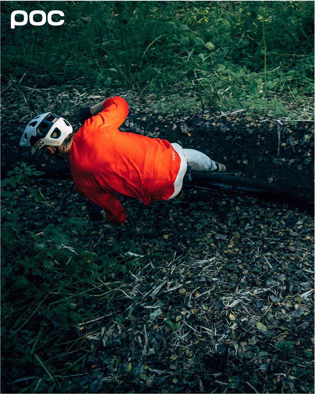 A cyclist in a red jacket and helmet rides through a forest trail, leaning into a turn on a dirt path surrounded by greenery. With Poc branding in the corner.