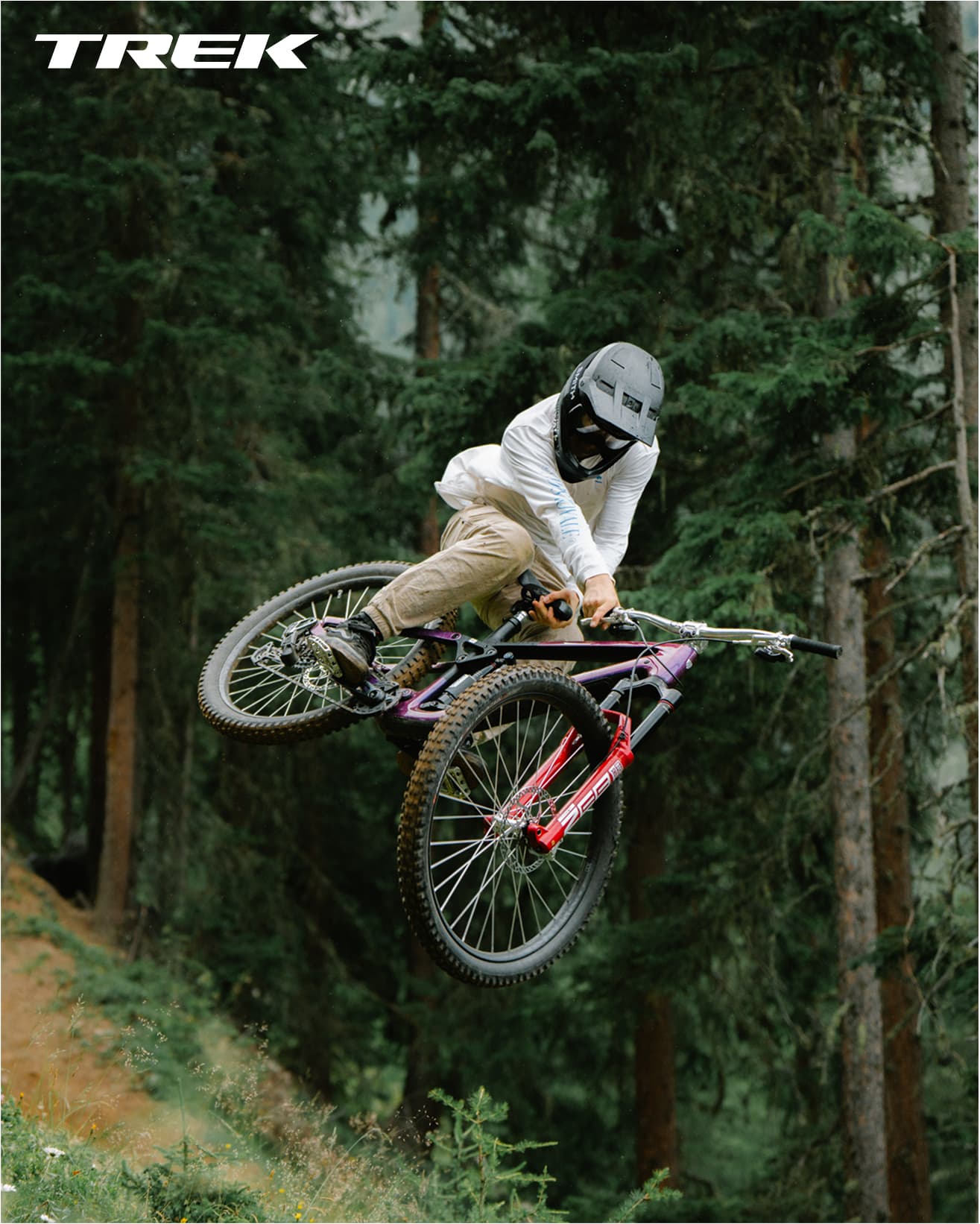Mountain biker in full gear performing a mid-air stunt on a trail in a forest, with Trek branding in the corner.
