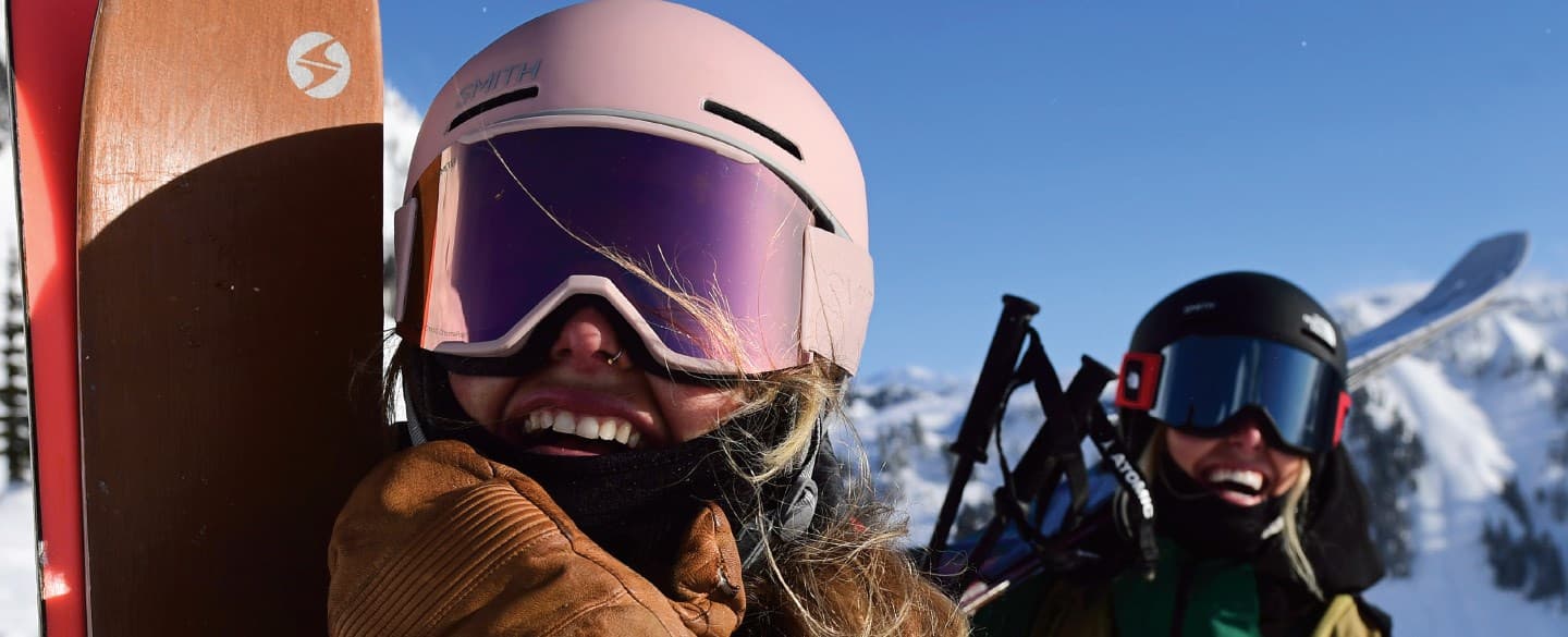 Two people in ski gear smiling, on a snowy mountain under a clear blue sky.