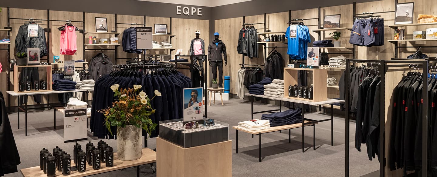 Clothing store interior with racks of shirts, jackets and shorts. Display tables hold folded clothes and accessories.