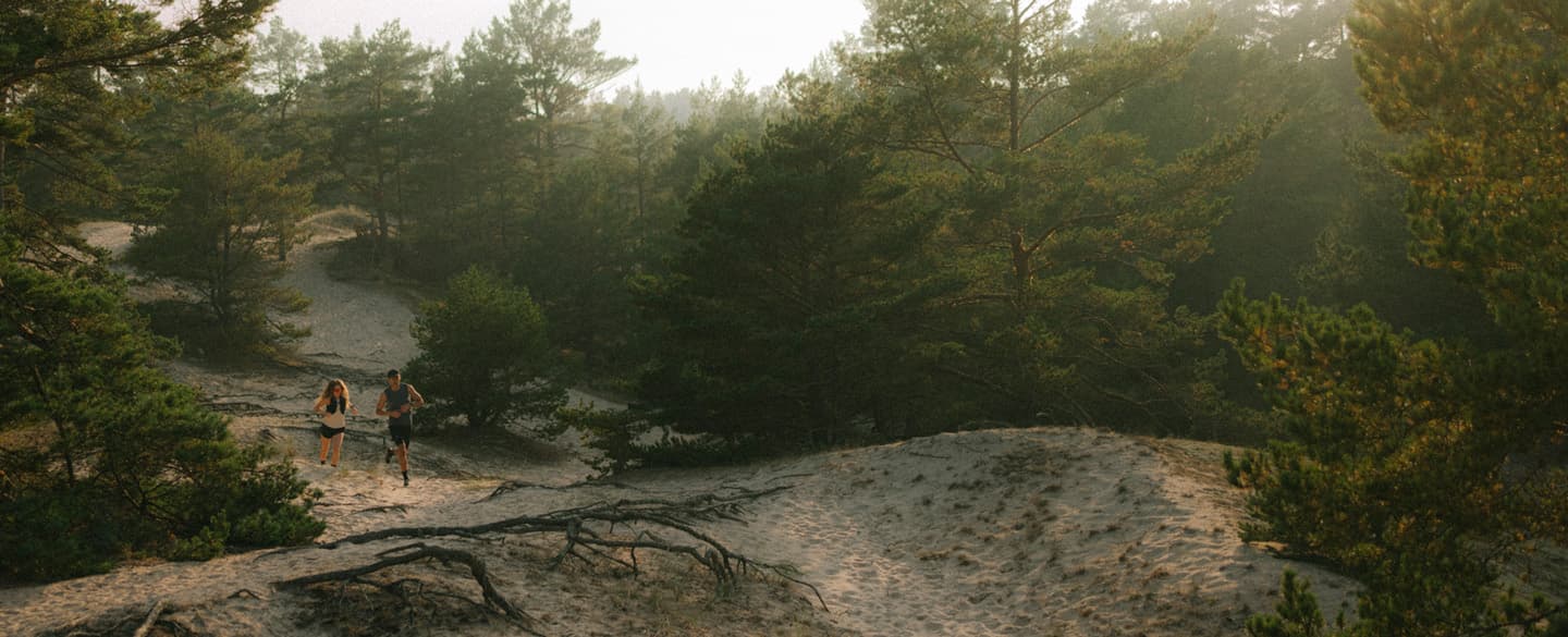 Two people running on a sandy path through a forested area with soft, morning light filtering through the trees.