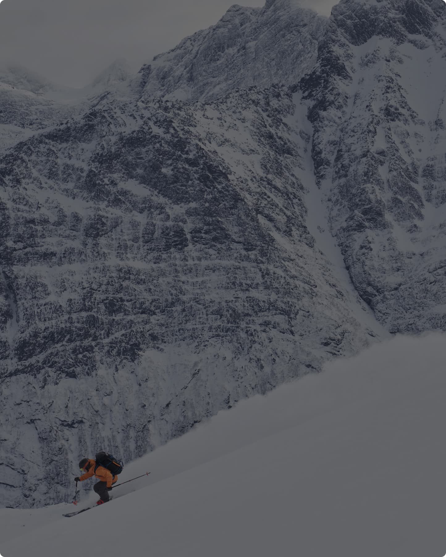 A skier in an orange jacket descends a snowy slope with large, rugged mountains in the background.