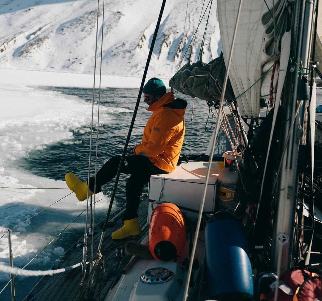 Person in yellow jacket and boots sits on sailboat deck beside sea ice, snowy mountains in background, ropes and gear visible on deck.
