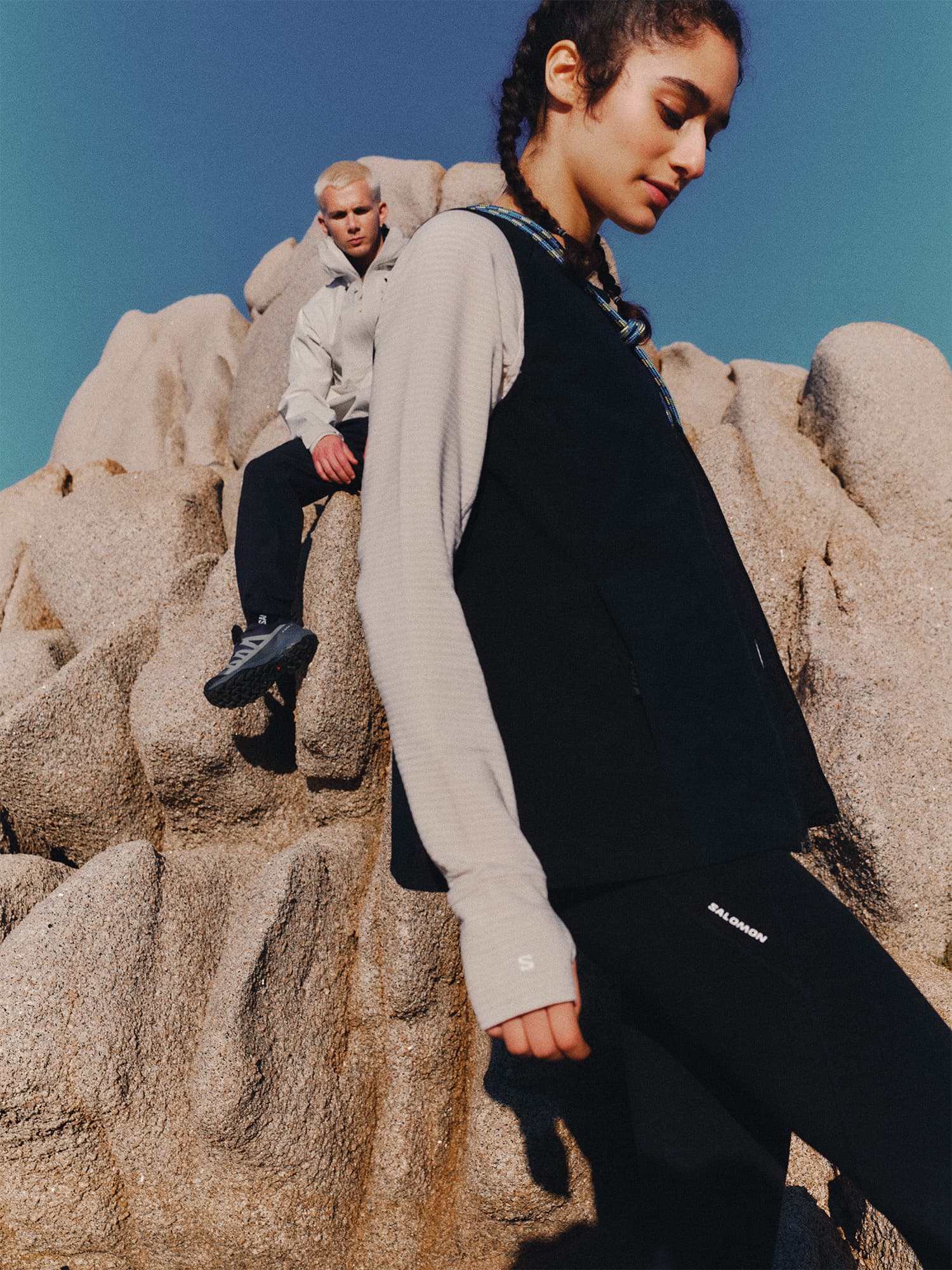 Two people in outdoor attire on rocky terrain. One sits on a rock, the other stands in the foreground. Clear blue sky above.