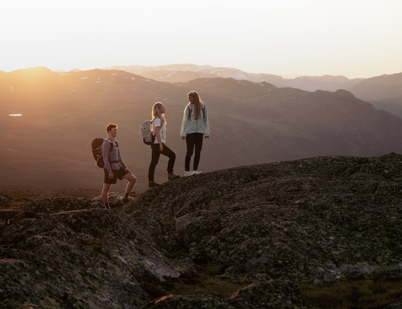 Three hikers with backpacks stand on rocky terrain, enjoying a sunrise view over distant mountains.