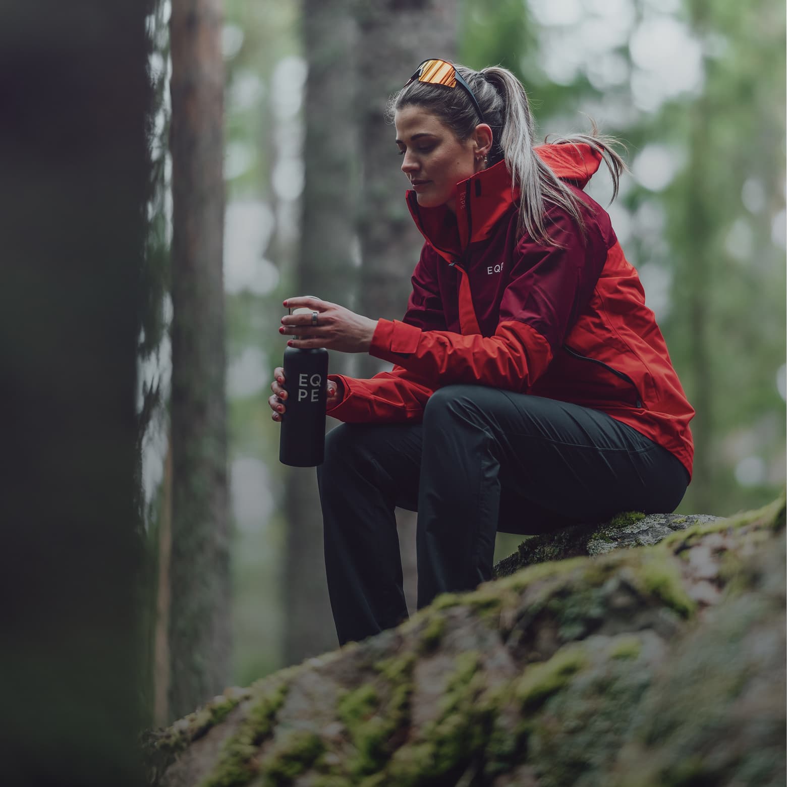 Woman wearing a red jacket, seated on a mossy rock in a forest, holding a black water bottle.