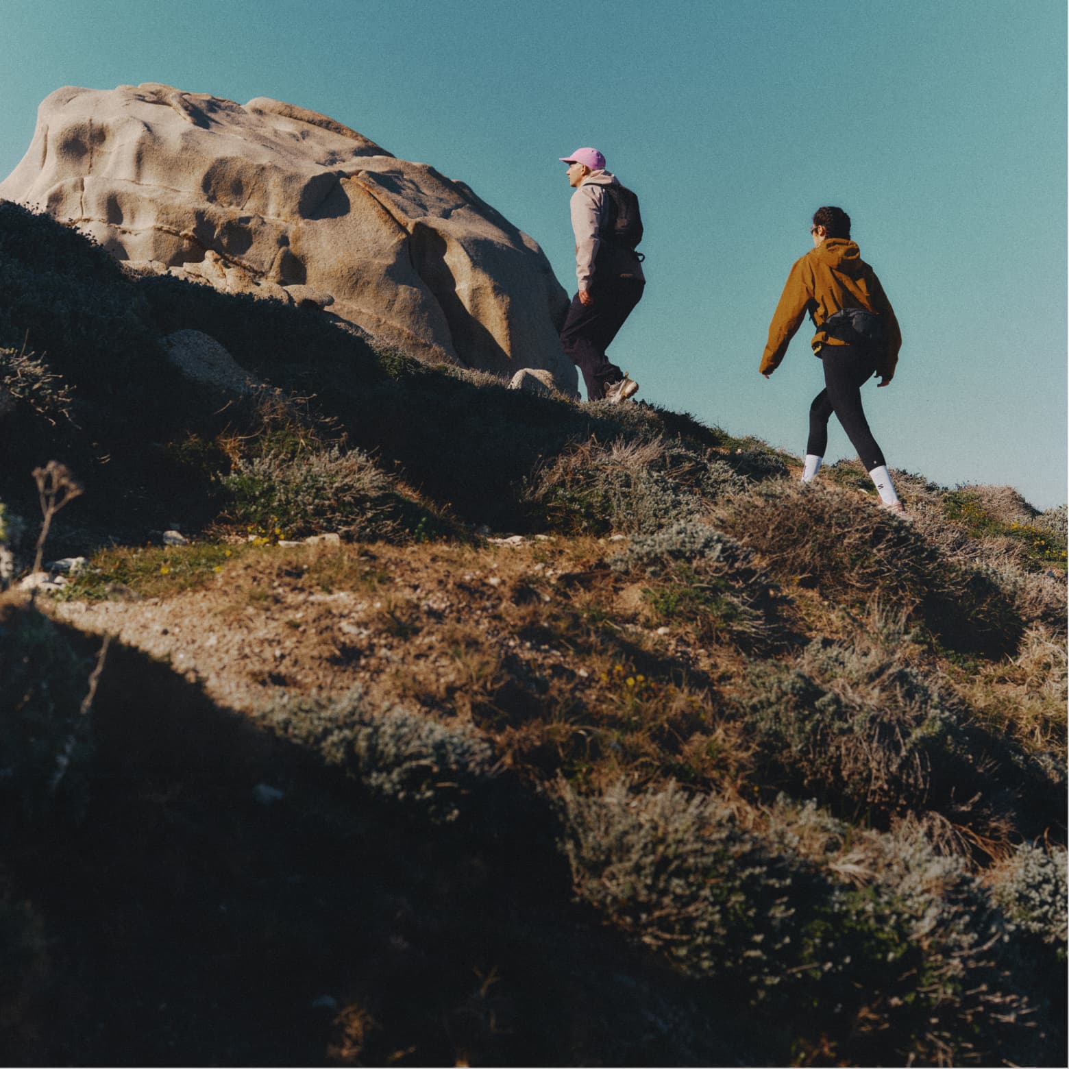 Two people hiking up a rocky hill with a large boulder in the background under a clear blue sky.