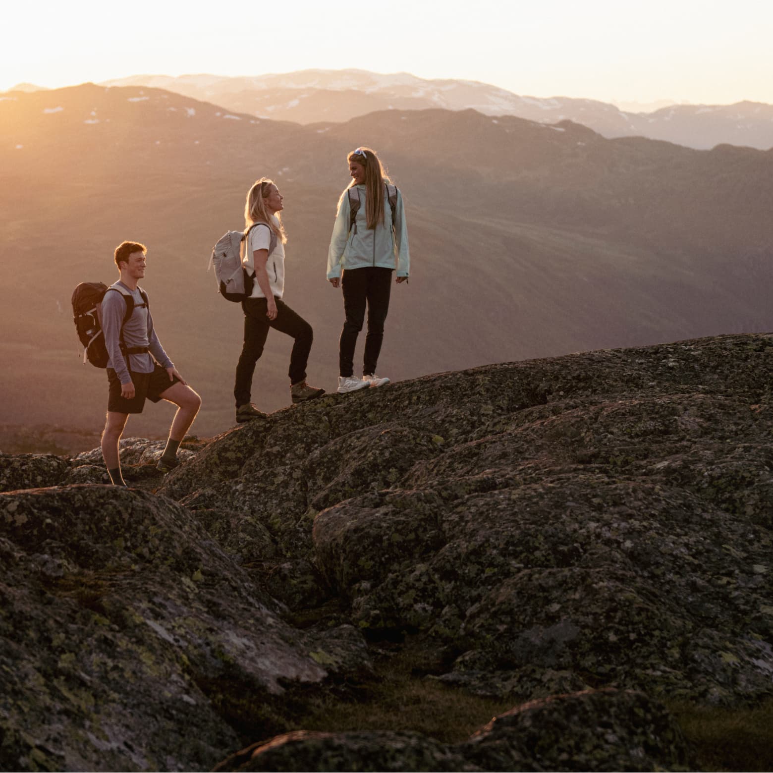 Three hikers with backpacks stand on rocky terrain, enjoying a sunrise view over distant mountains.