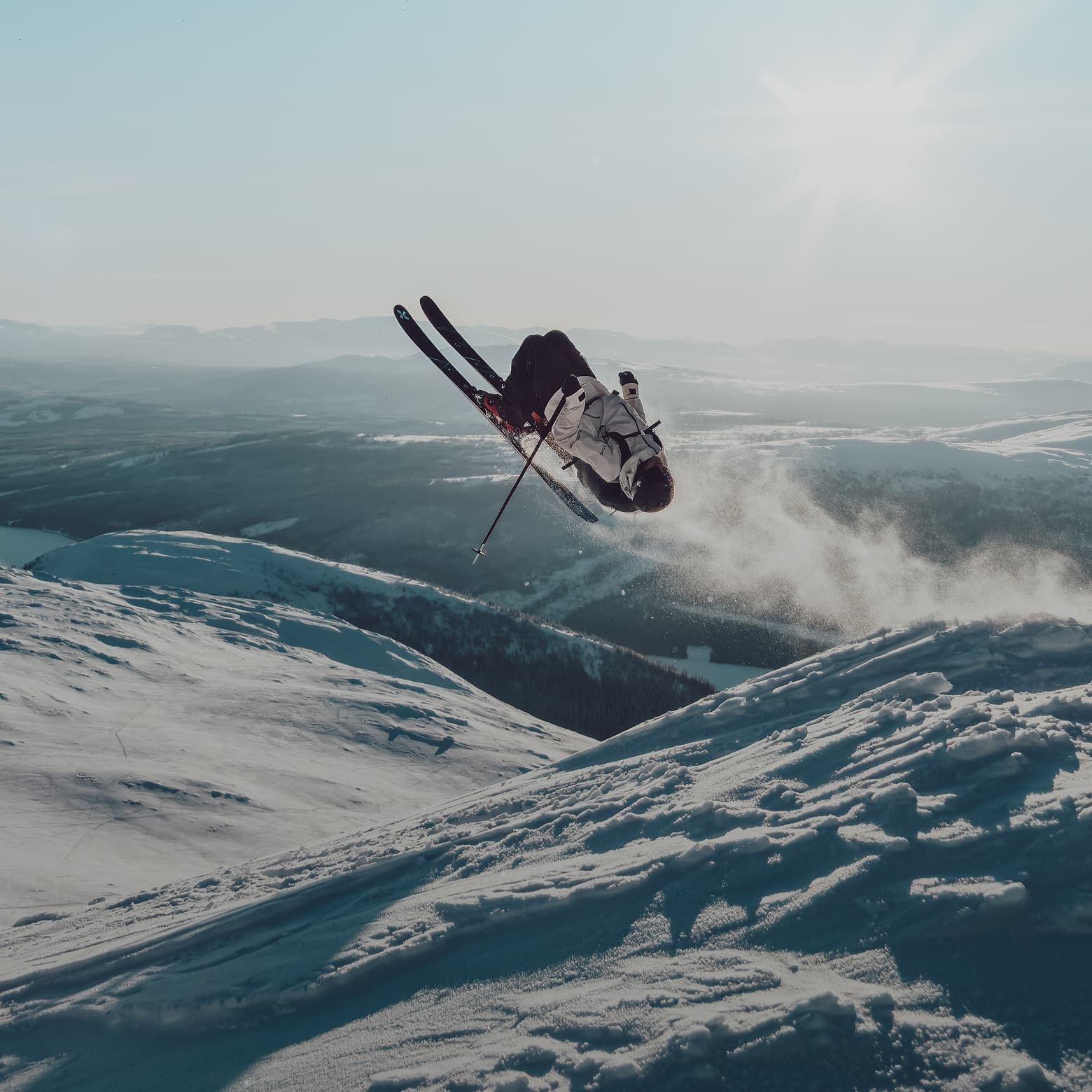 A skier performs a mid-air flip against a backdrop of snowy mountains and expansive landscape under a cloudy sky.