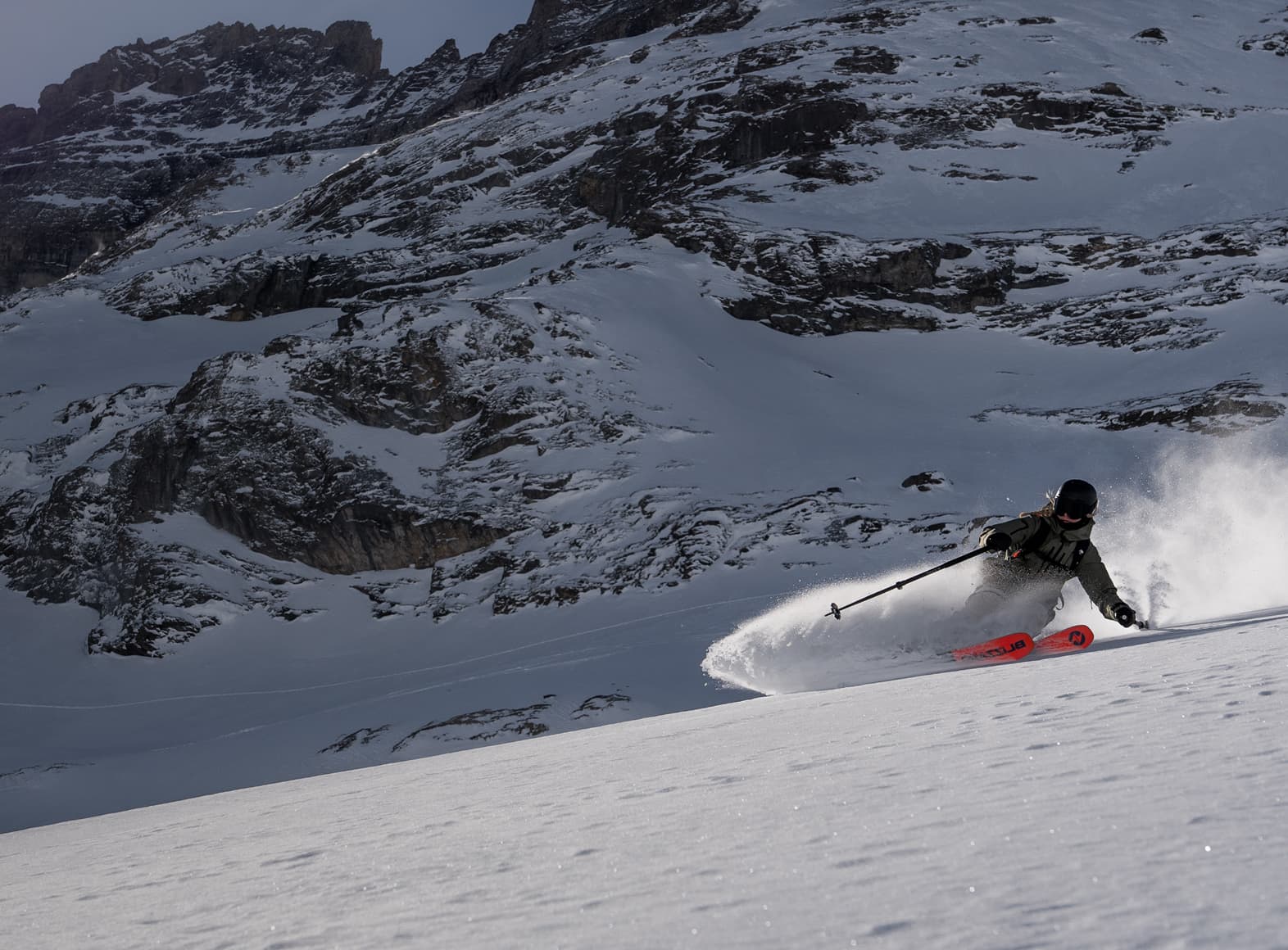 A skier in green gear and red skis carves through fresh snow on a steep, snowy mountain slope under a clear sky.