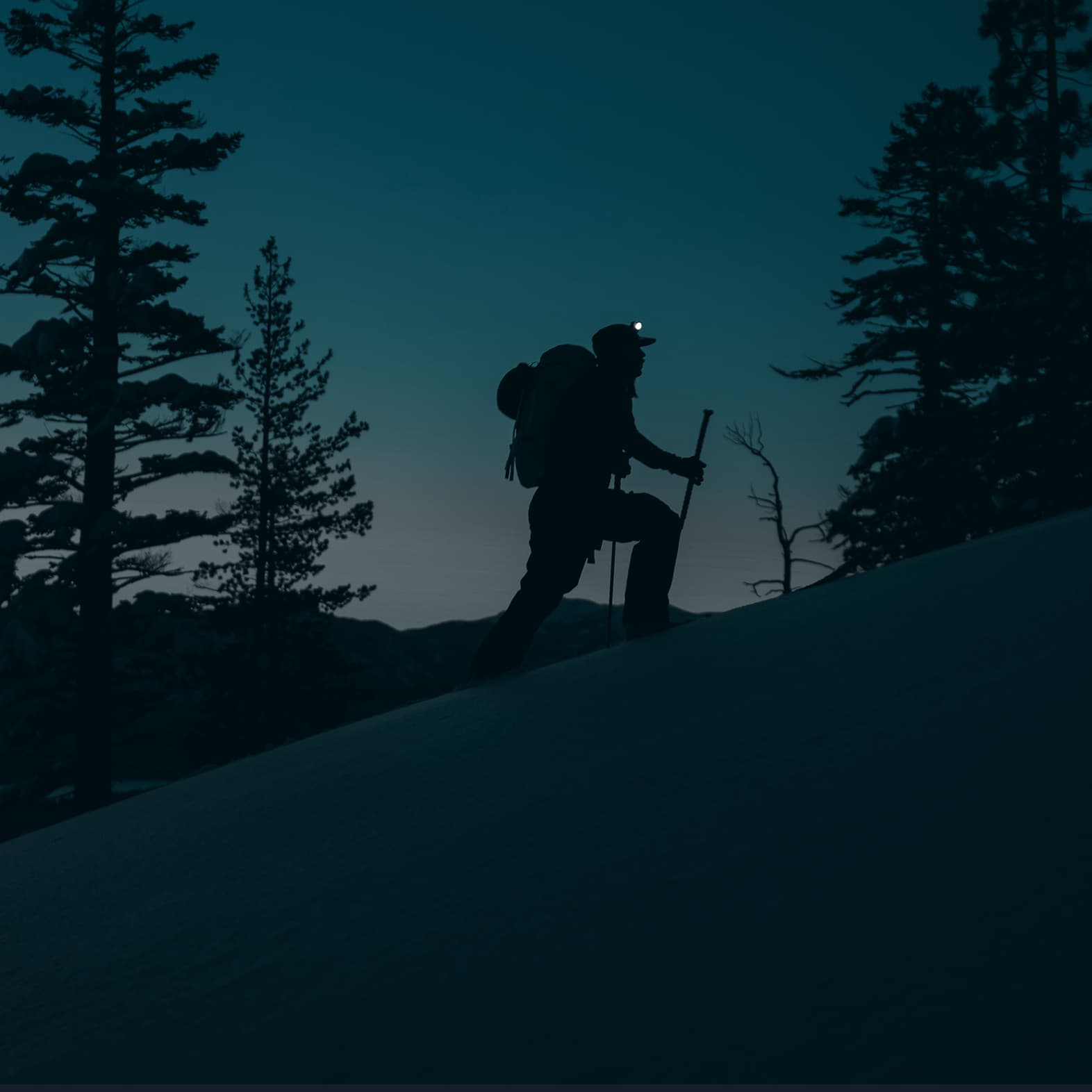 Silhouette of a hiker with a headlamp and backpack, climbing a snowy hill at dusk, surrounded by trees.