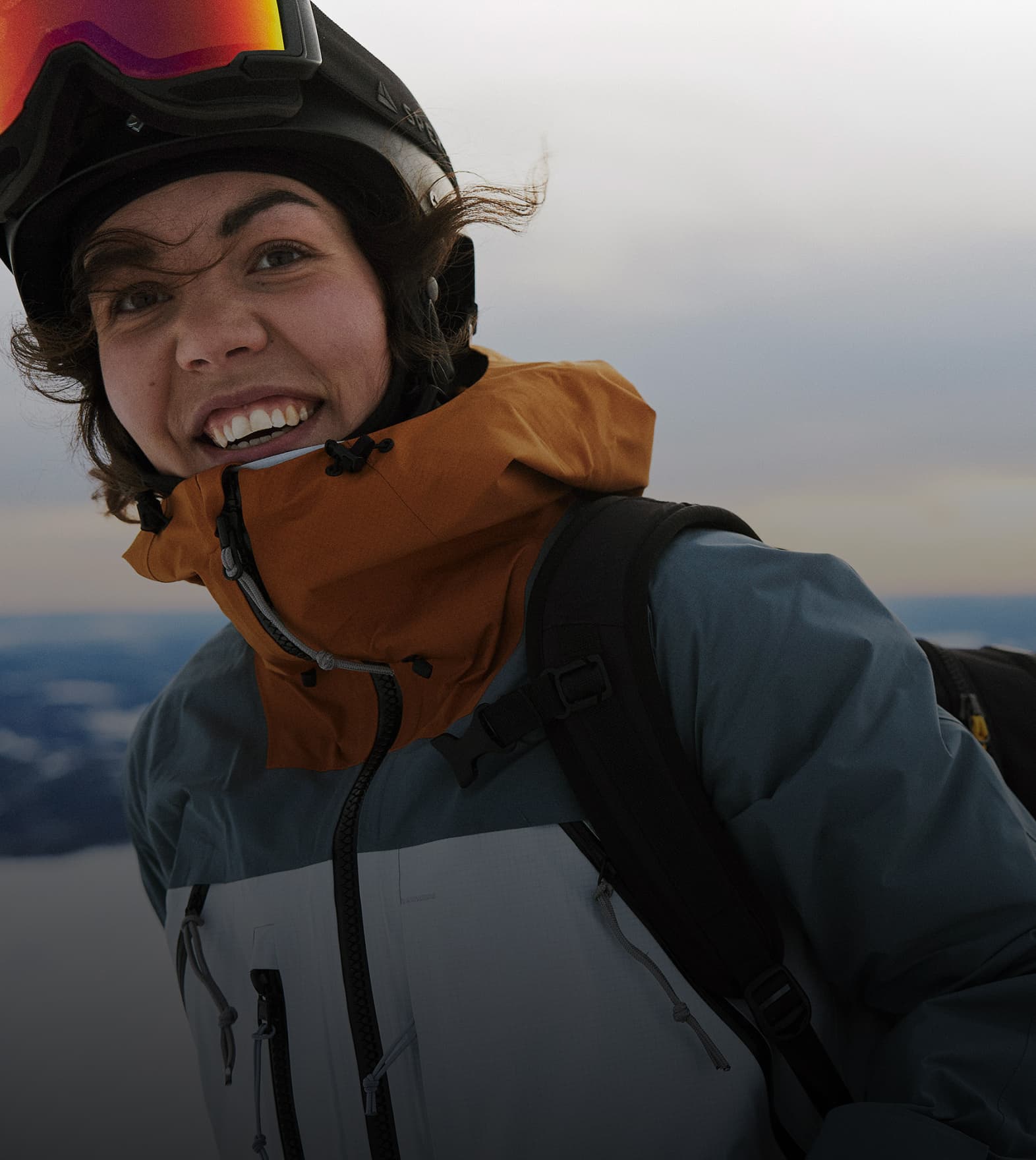 Person in ski gear smiling, with a snowy landscape and cloudy sky in the background.