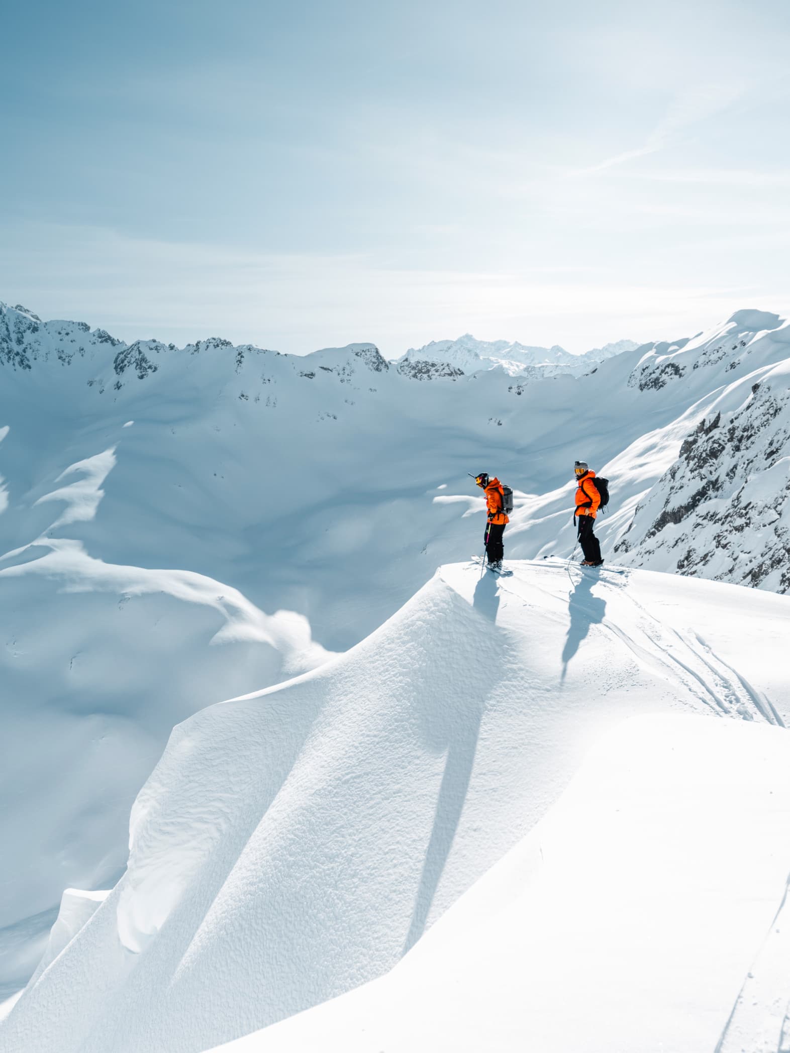 Two skiers in orange jackets stand on a snowy mountain peak, overlooking a vast, snow-covered landscape under a clear blue sky.