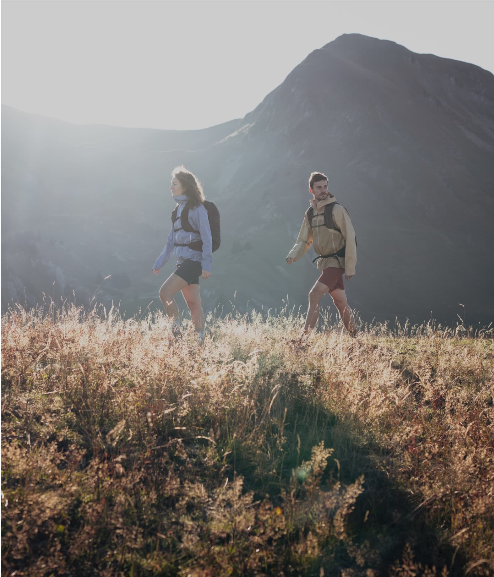Two people hiking on a grassy trail with sunlight and mountains in the background.