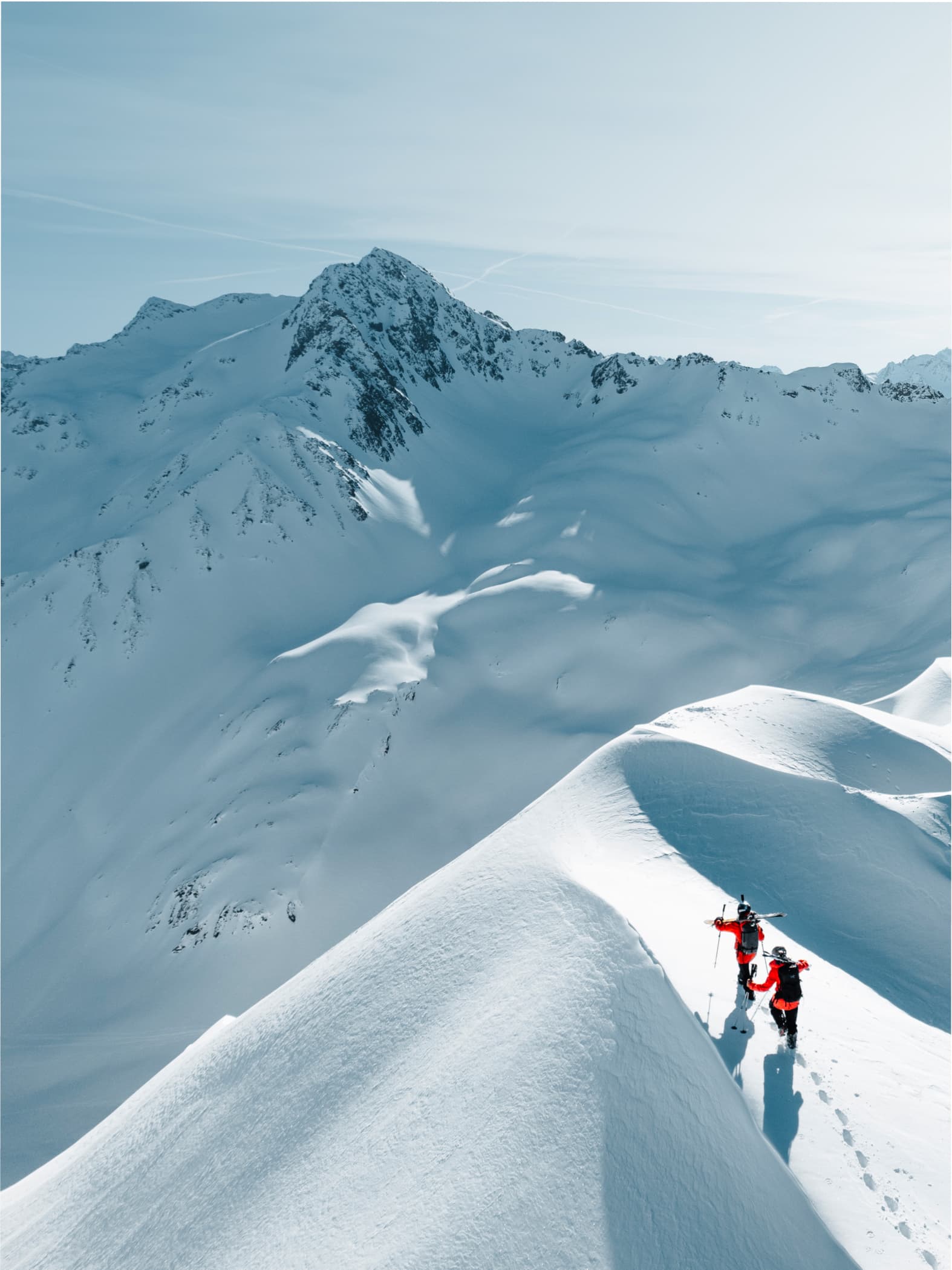 Two skiers in red jackets traverse a snowy mountain ridge under a clear blue sky, leaving footprints in the pristine snow.