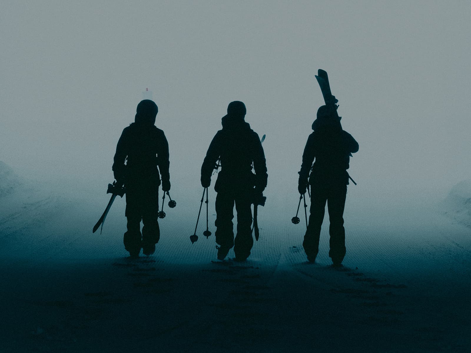 Three silhouetted figures in winter gear walk across a snowy, foggy landscape carrying poles and packs.