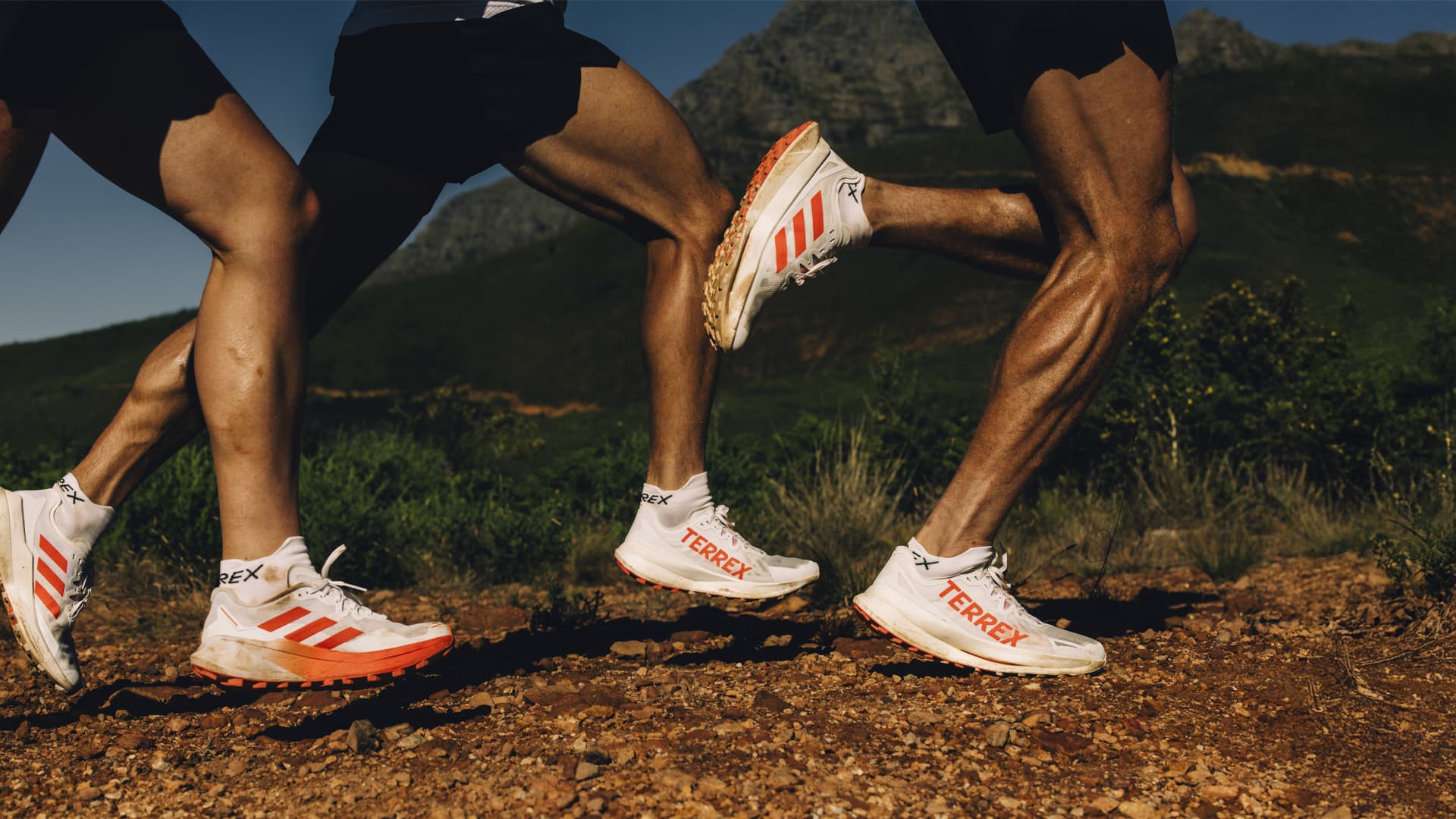 Three runners on a rocky trail, wearing white and orange running shoes, with mountains in the background.