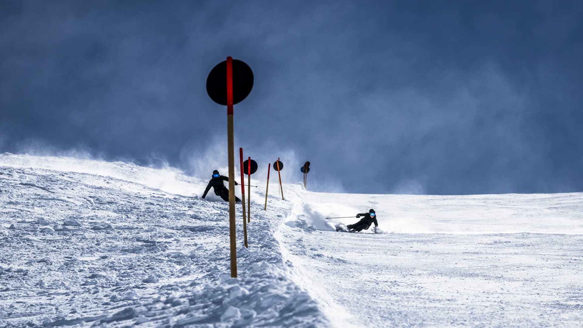 Two skiers in dark outfits and blue reflective goggles carve down a snowy slope marked with red-tipped poles. Snow sprays behind the skiers as they make sharp turns.