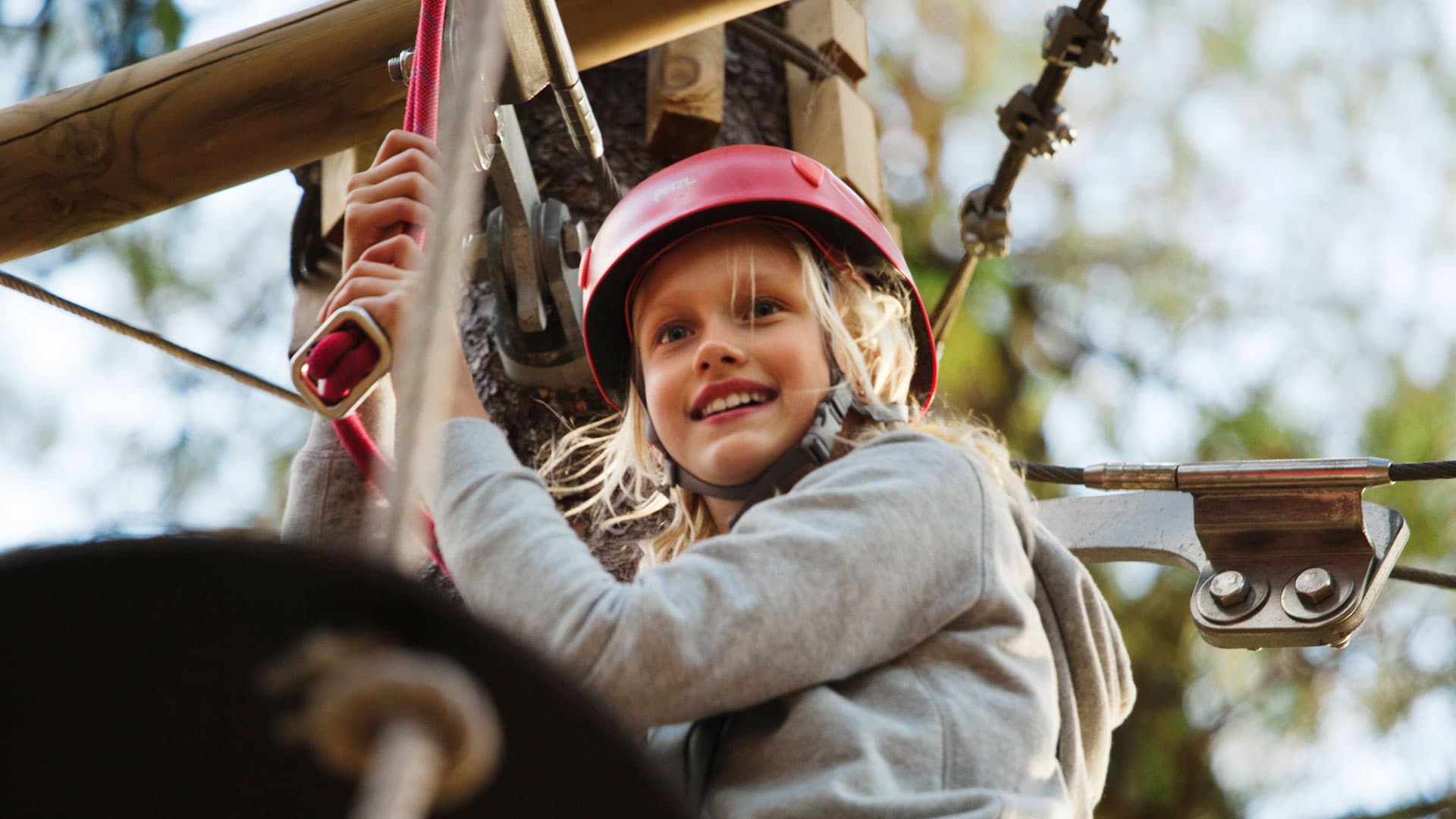 A smiling child wearing a red helmet is climbing a rope course, gripping a red rope, surrounded by trees.