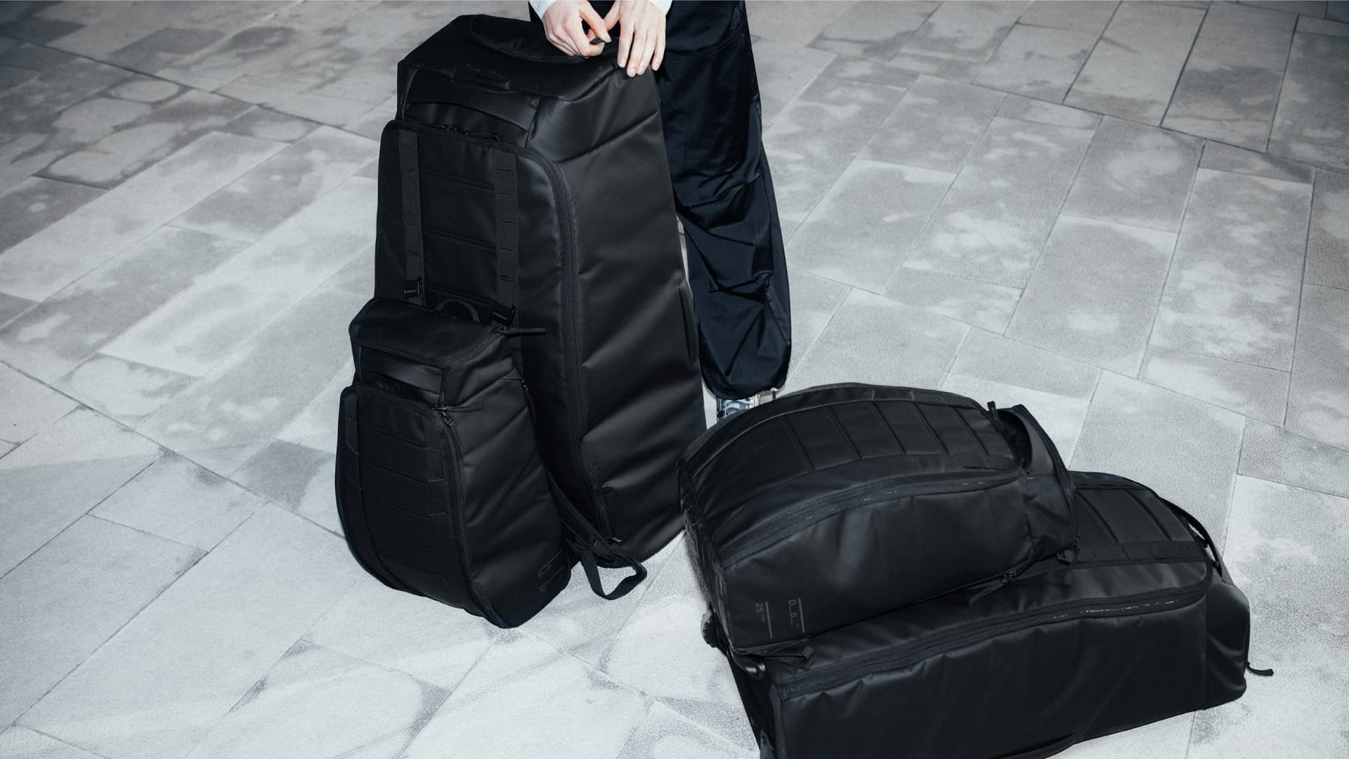 Person standing on pavement with three large black travel bags, including a backpack and duffel, arranged on the ground.