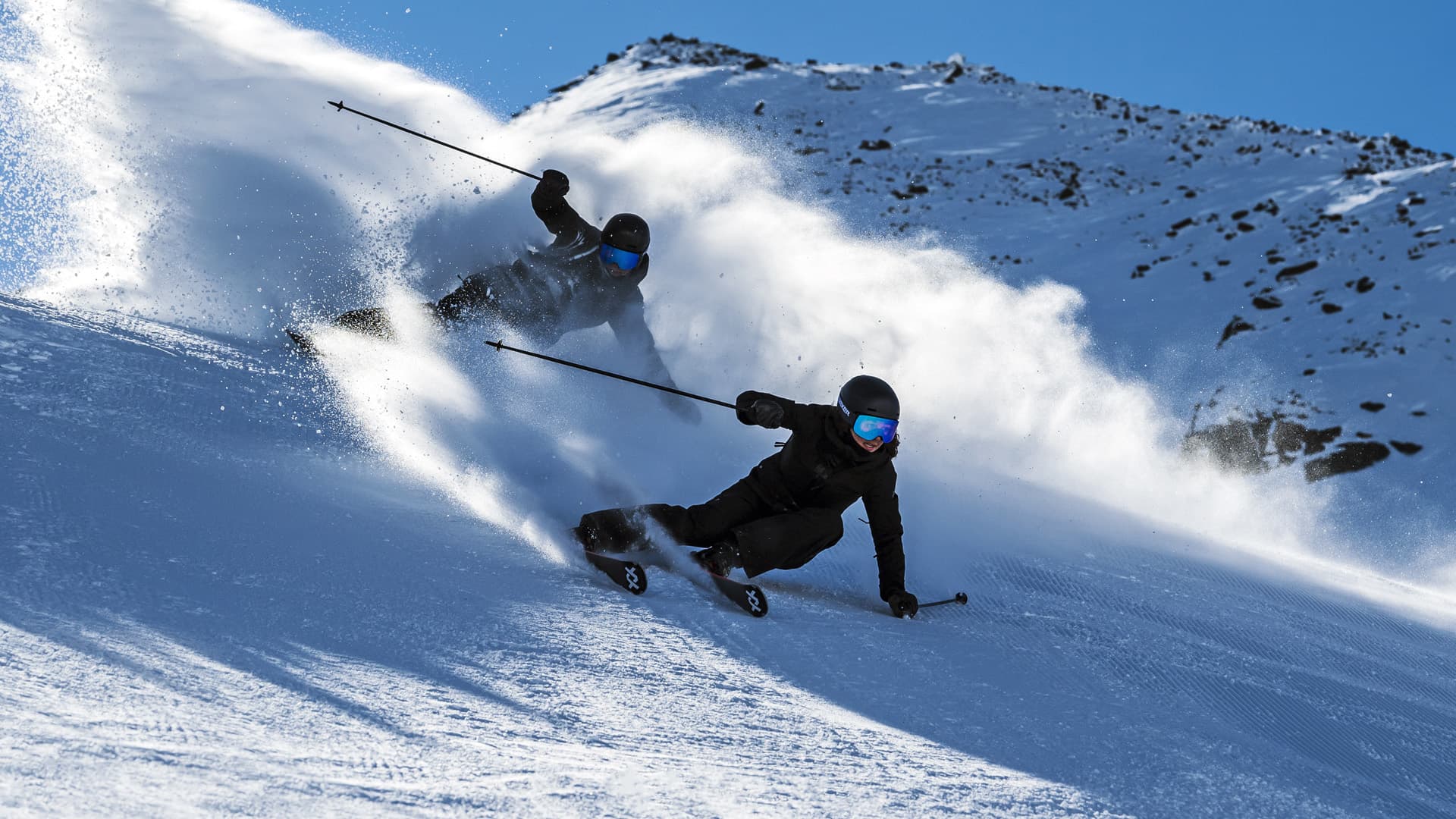 Two skiers in black gear carving down a snowy slope, creating trails of powder under a clear blue sky with rocky peaks in the background.