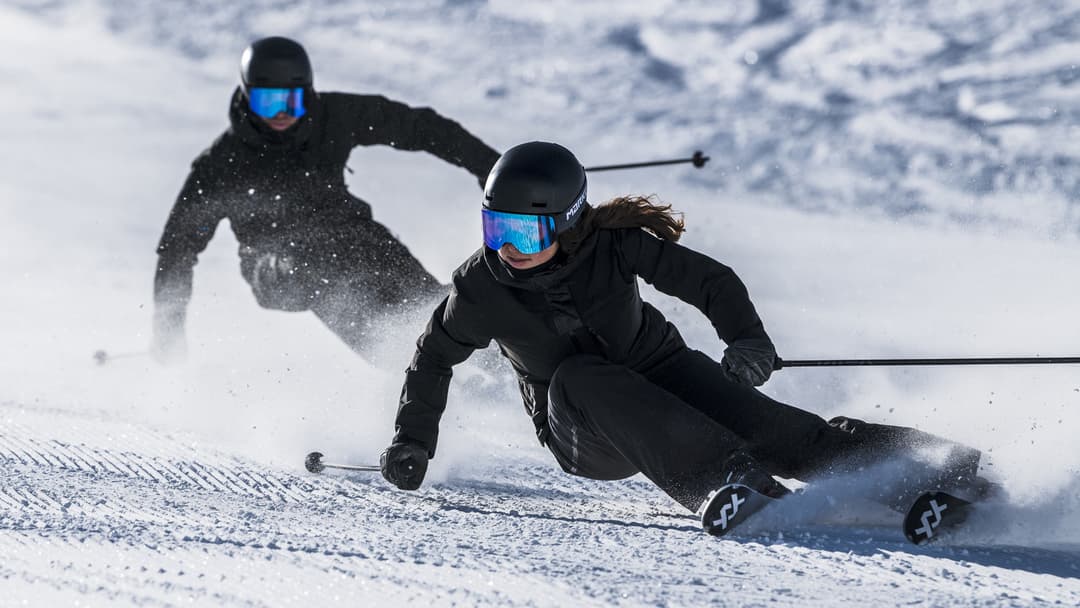 Two skiers in black outfits and reflective ski goggles carve swiftly down a snowy slope, leaving trails of powder behind them.