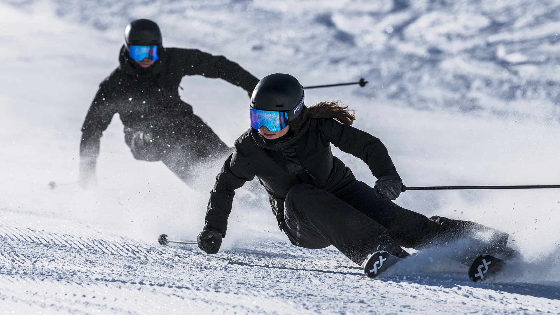 Two skiers in black outfits and reflective ski goggles carve swiftly down a snowy slope, leaving trails of powder behind them.